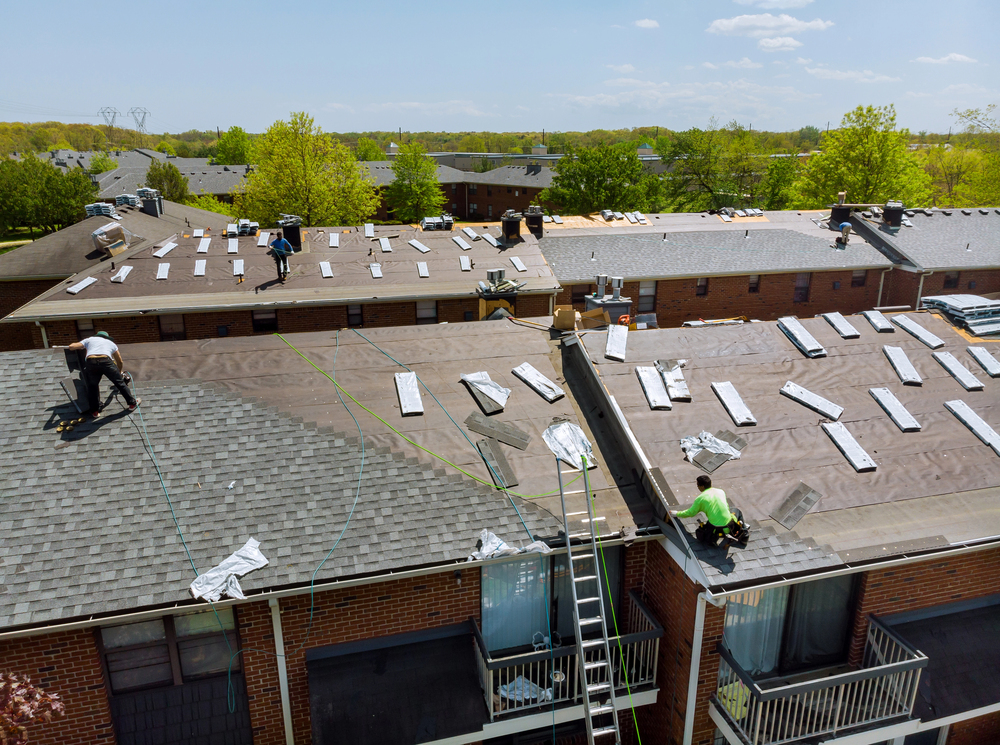 Workers installing new roofing materials on a large apartment building; shingles and tools are spread across the roof under clear skies.