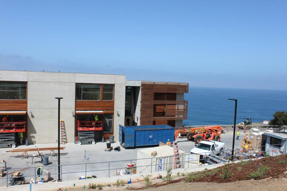 A modern building under construction near the ocean, with construction vehicles, workers, and equipment visible in the foreground.