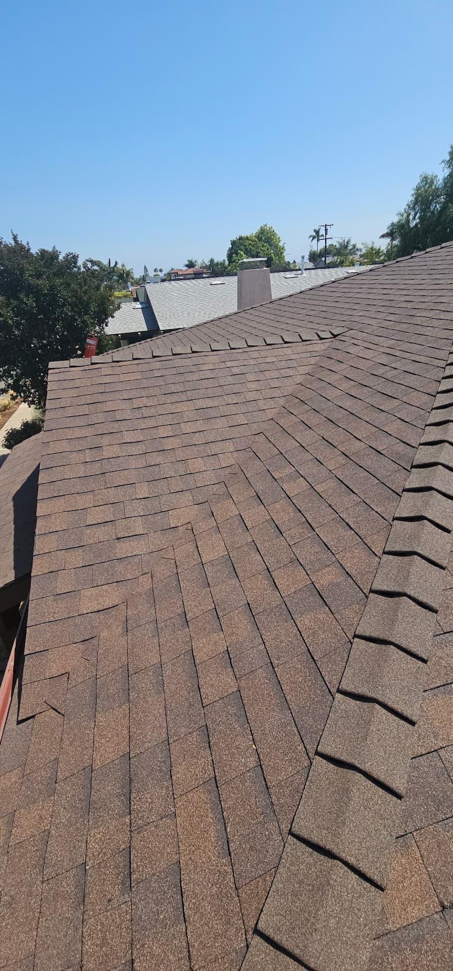 A view of a residential roof with brown asphalt shingles after a recent roof replacement, under a clear blue sky, with trees and rooftops visible in the background.