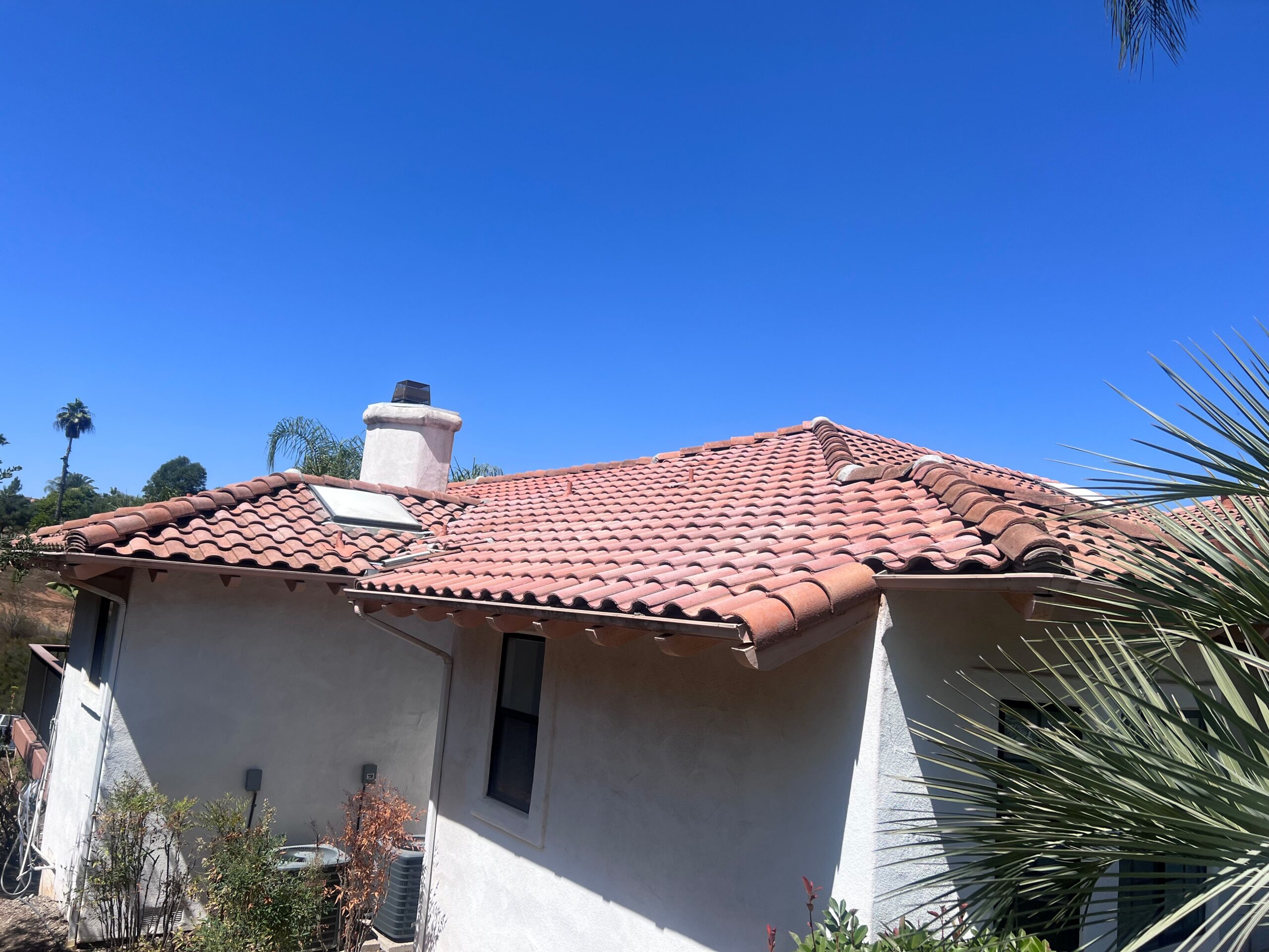 A single-story house with red tile roofing, a chimney, and a skylight—ideal for easy roof repair—set against a clear blue sky with some plants in the foreground.