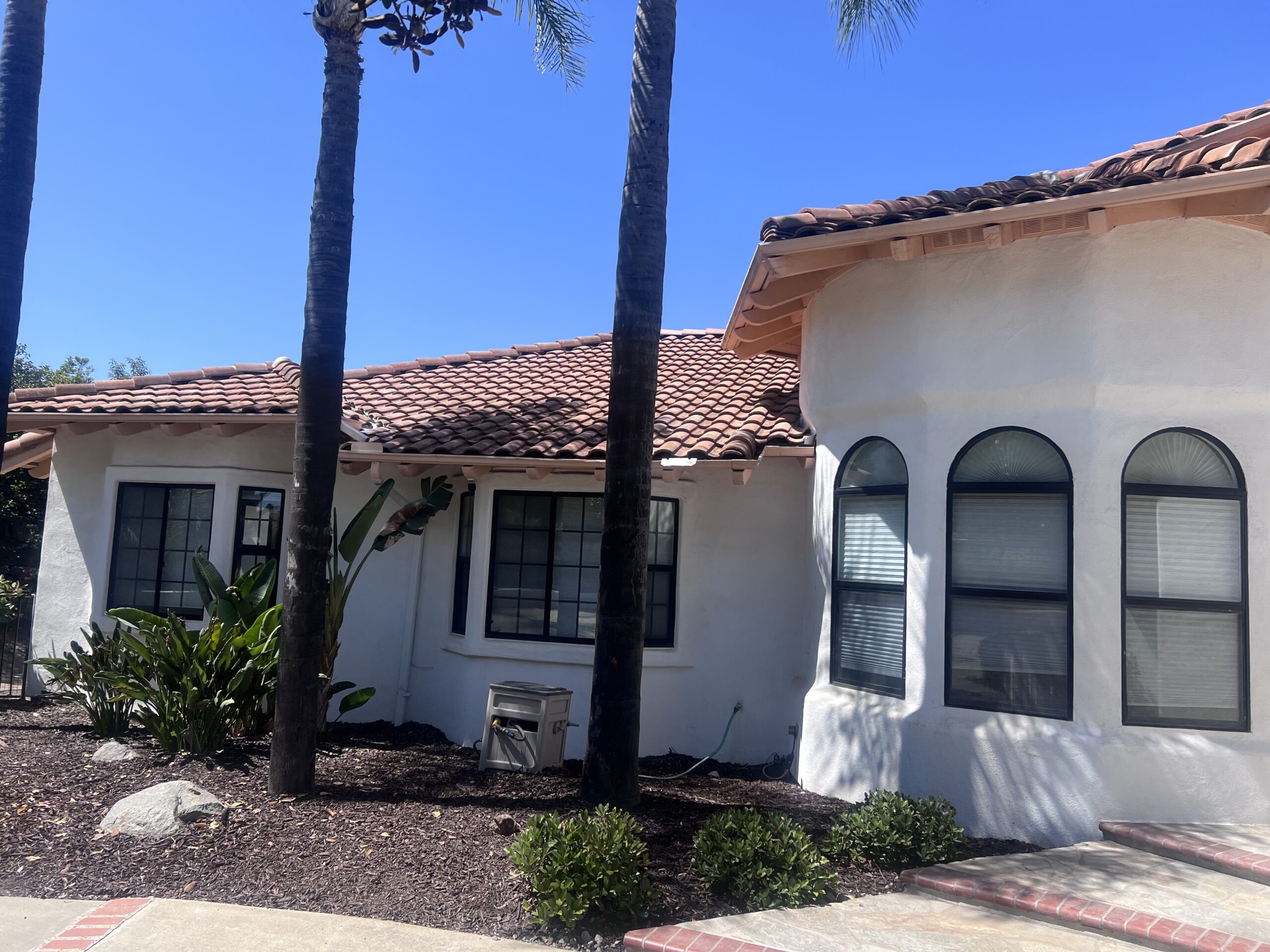Single-story white stucco house with a red tile roof covered by a roofing warranty, black-trimmed windows, and an arched window section, surrounded by palm trees and small shrubs.